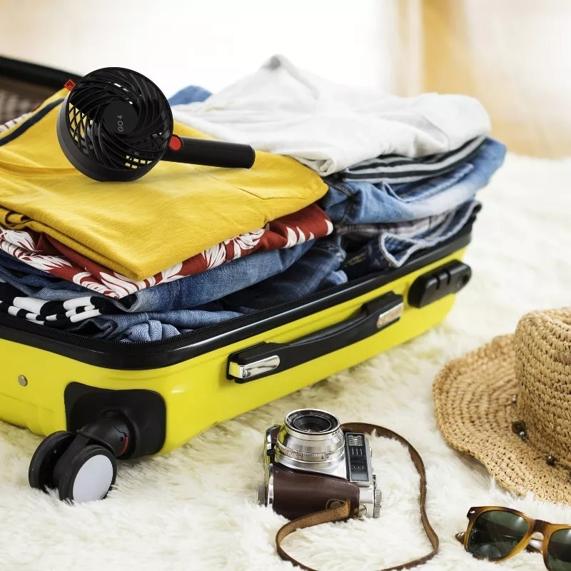 Open suitcase with clothes, hairdryer, camera, and headphones next to a hat and sunglasses, suggesting travel essentials