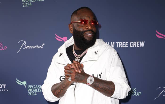 Rick Ross in a white jacket with accessories, posing at an event