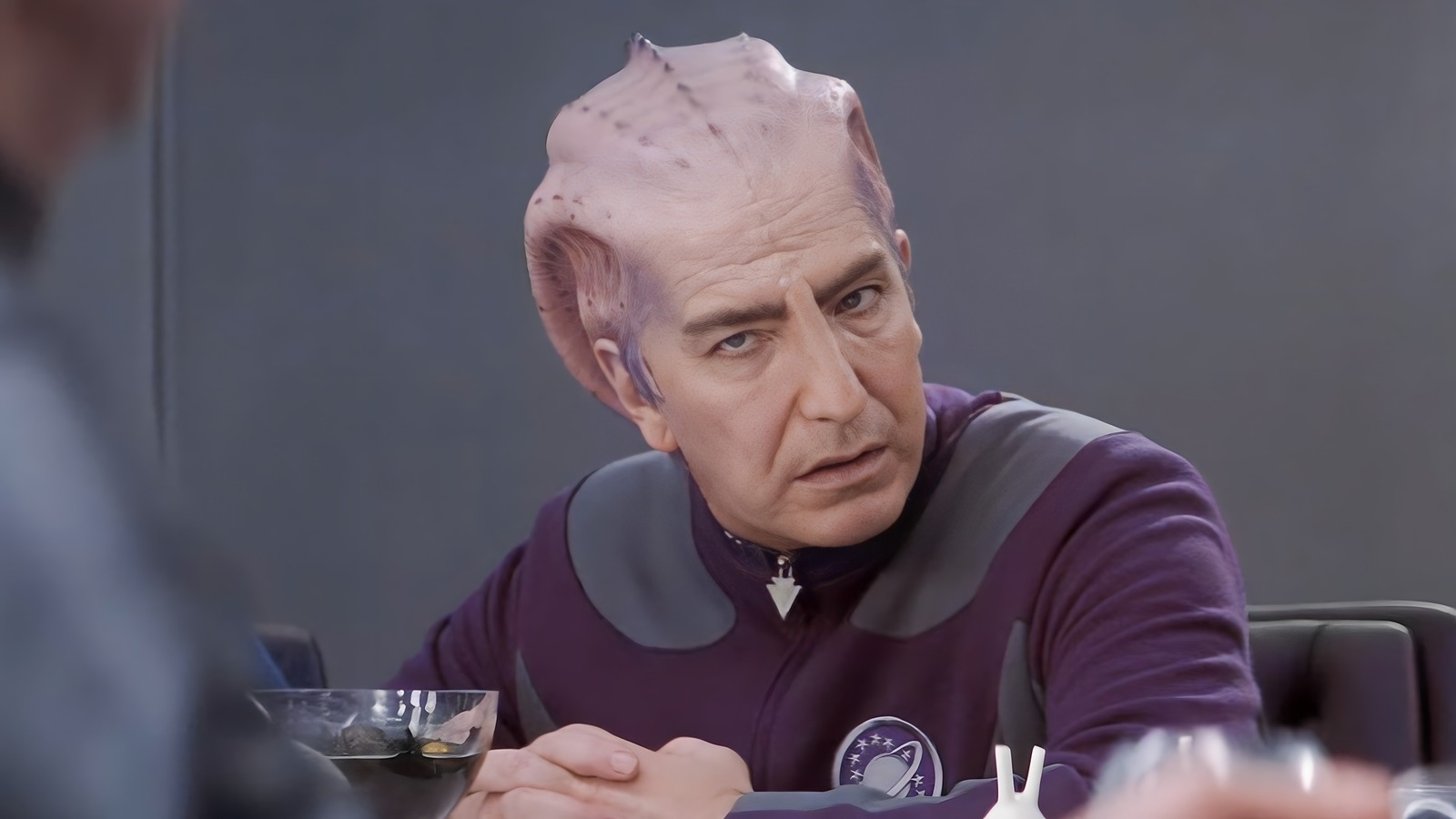 Character in Starfleet uniform with alien prosthetics looking pensive at a meeting