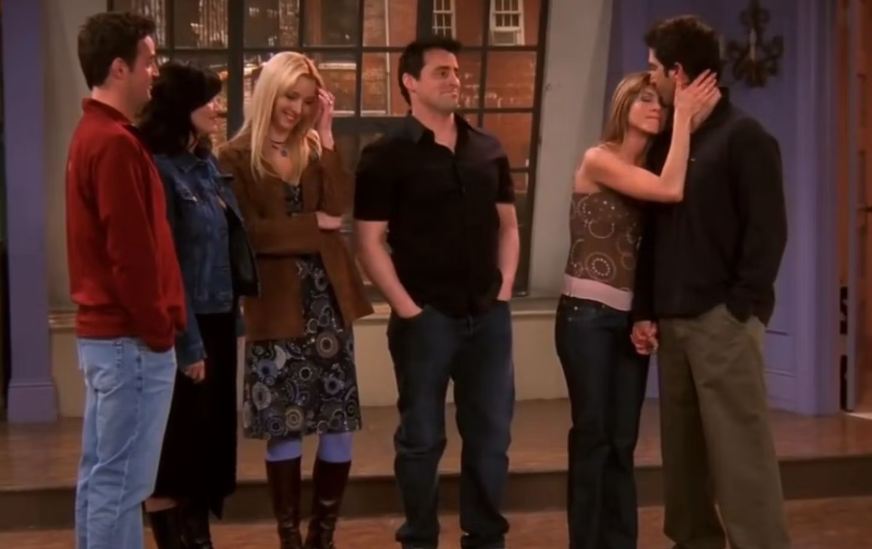 Group of people gathered, two are kissing with others watching; from TV show