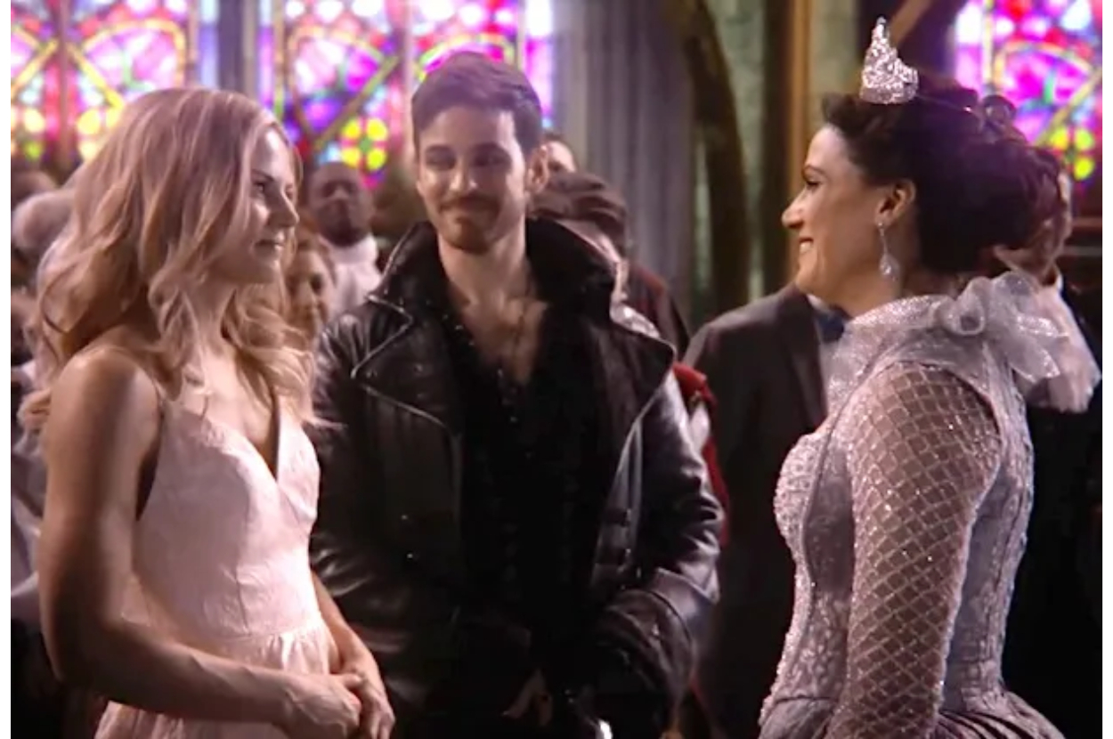 Three characters from a film or show, two women in dresses with one wearing a tiara and a man in a leather jacket, interacting indoors