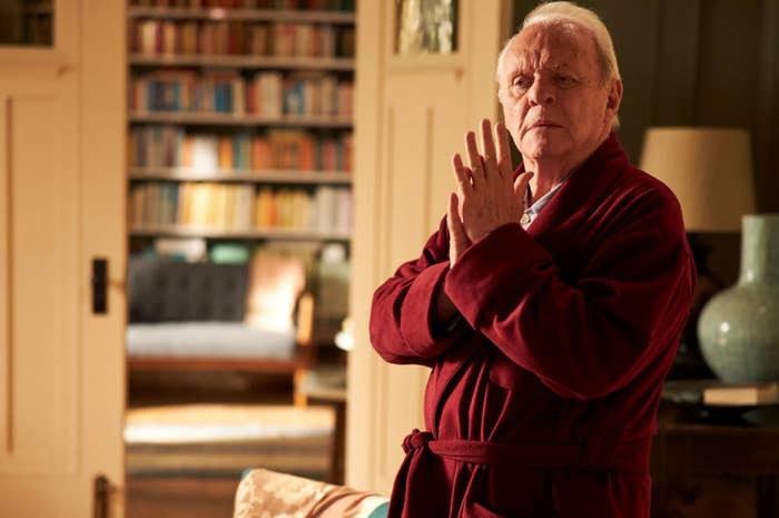 Sir Anthony Hopkins in a red robe, with hands clasped, standing indoors near a bookshelf