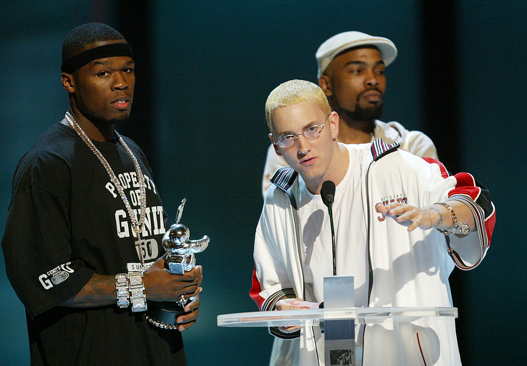 Three individuals at an awards ceremony, center person speaking at a microphone, all dressed in hip-hop style attire