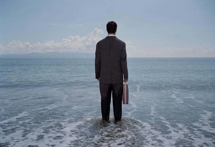 Person in a suit standing in the sea with a briefcase, facing away from the camera