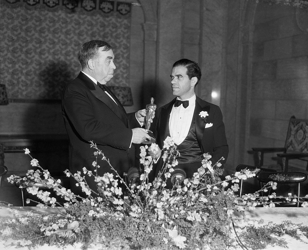Two men at an award ceremony, one in a tuxedo receiving a trophy from the other in a suit
