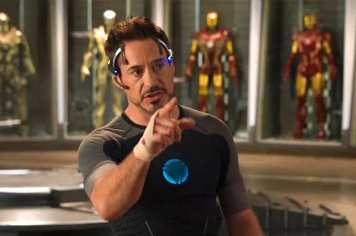 Tony Stark in a workshop with Iron Man suits in the background, pointing forward