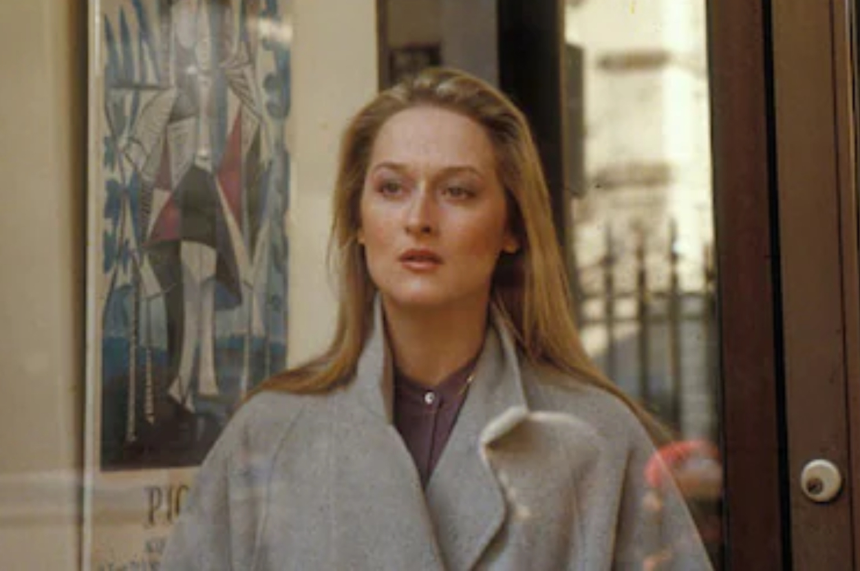 Meryl Streep in a grey coat and purple top, looking thoughtful in a movie scene