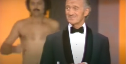 Man in a tuxedo stands at a podium while another shirtless man appears behind him on stage