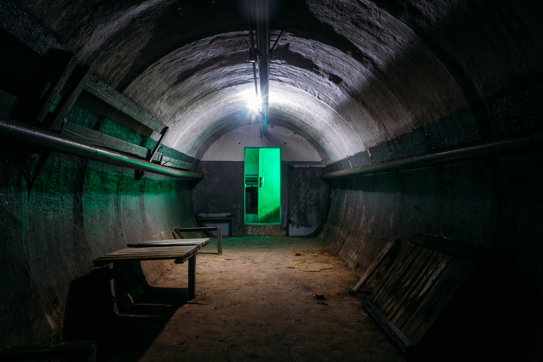 An illuminated green door at the end of a dark, arch-shaped tunnel with benches on sides
