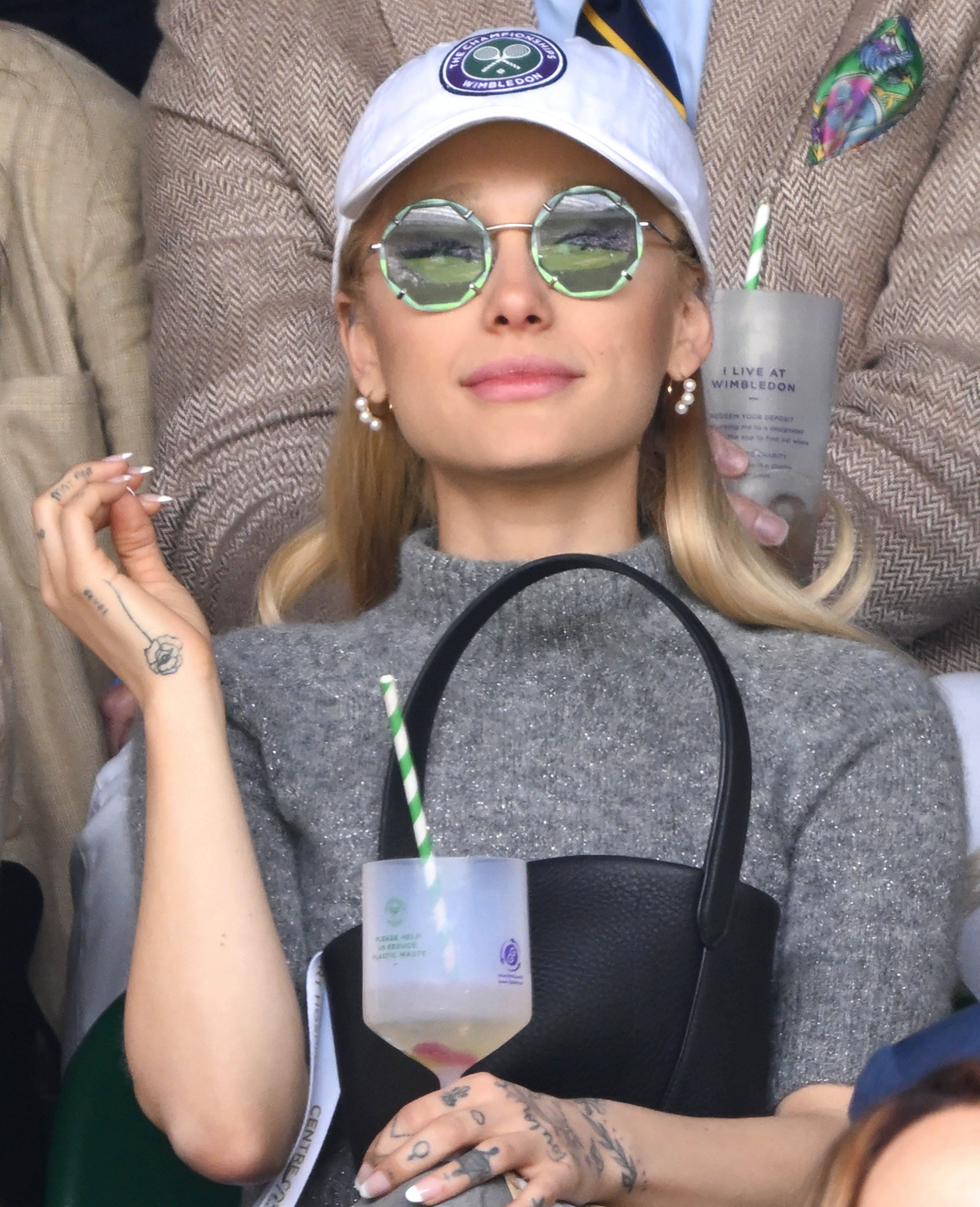 Ariana wears a baseball cap and sunglasses at an event and holds a drink