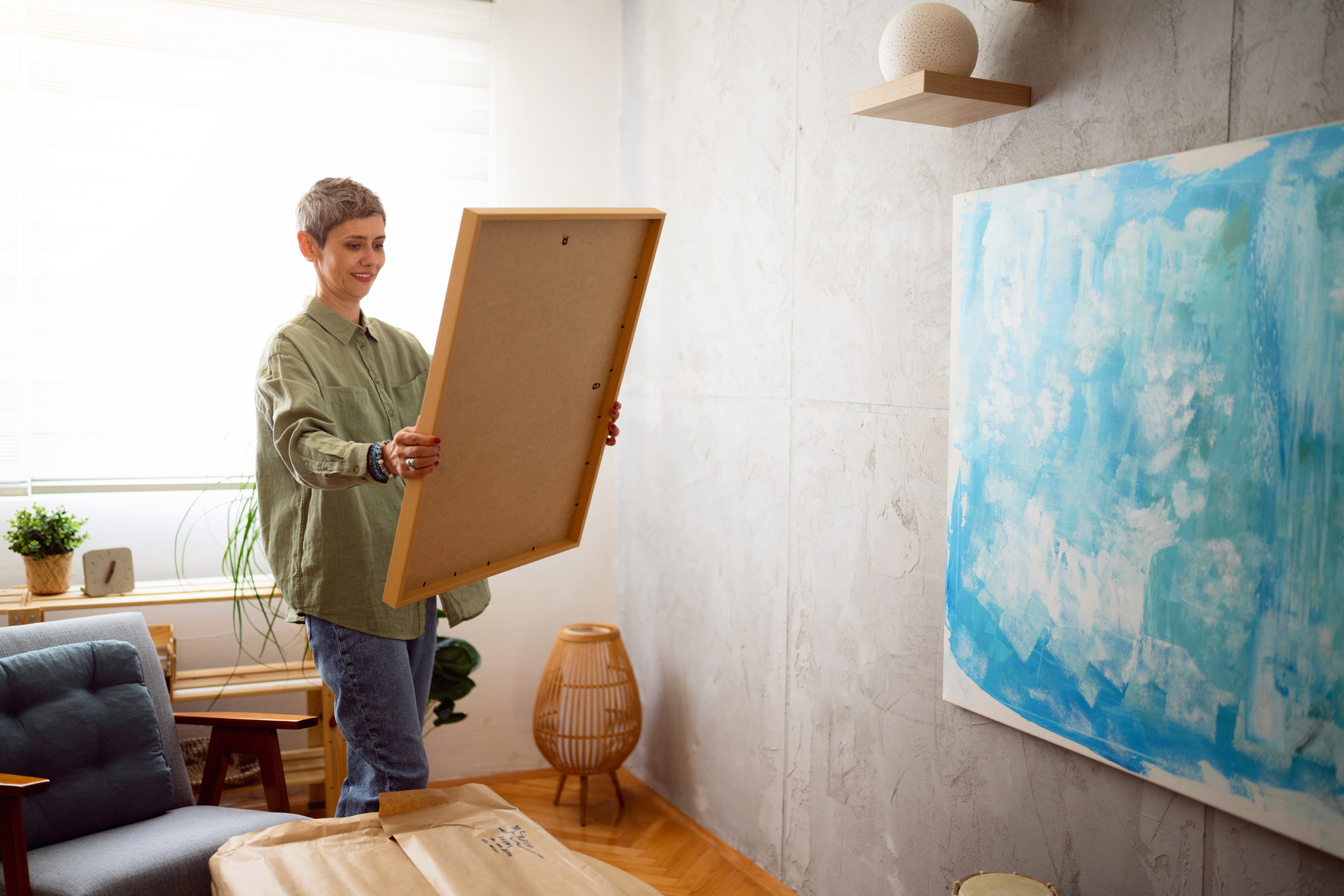 Person hanging a picture frame in a room with furniture and a large blue painting on the wall