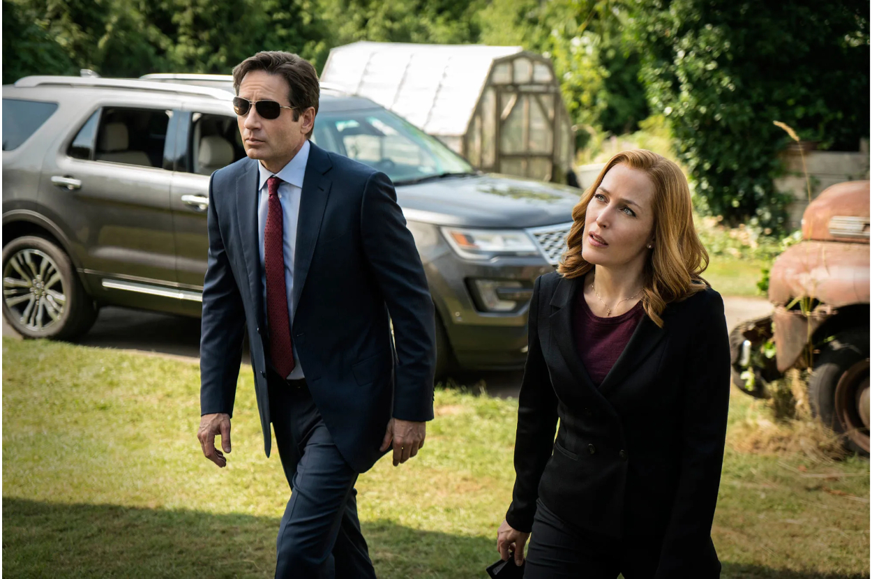 Two actors portraying characters walking, the man in a suit and sunglasses, the woman in a jacket and blouse