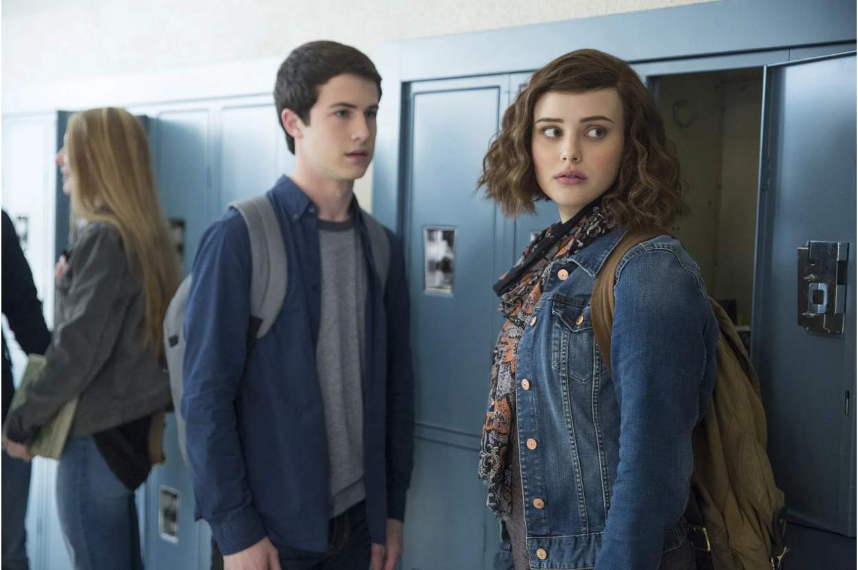 Two characters from a TV show stand near lockers, one with a backpack, the other wearing a denim jacket with patches