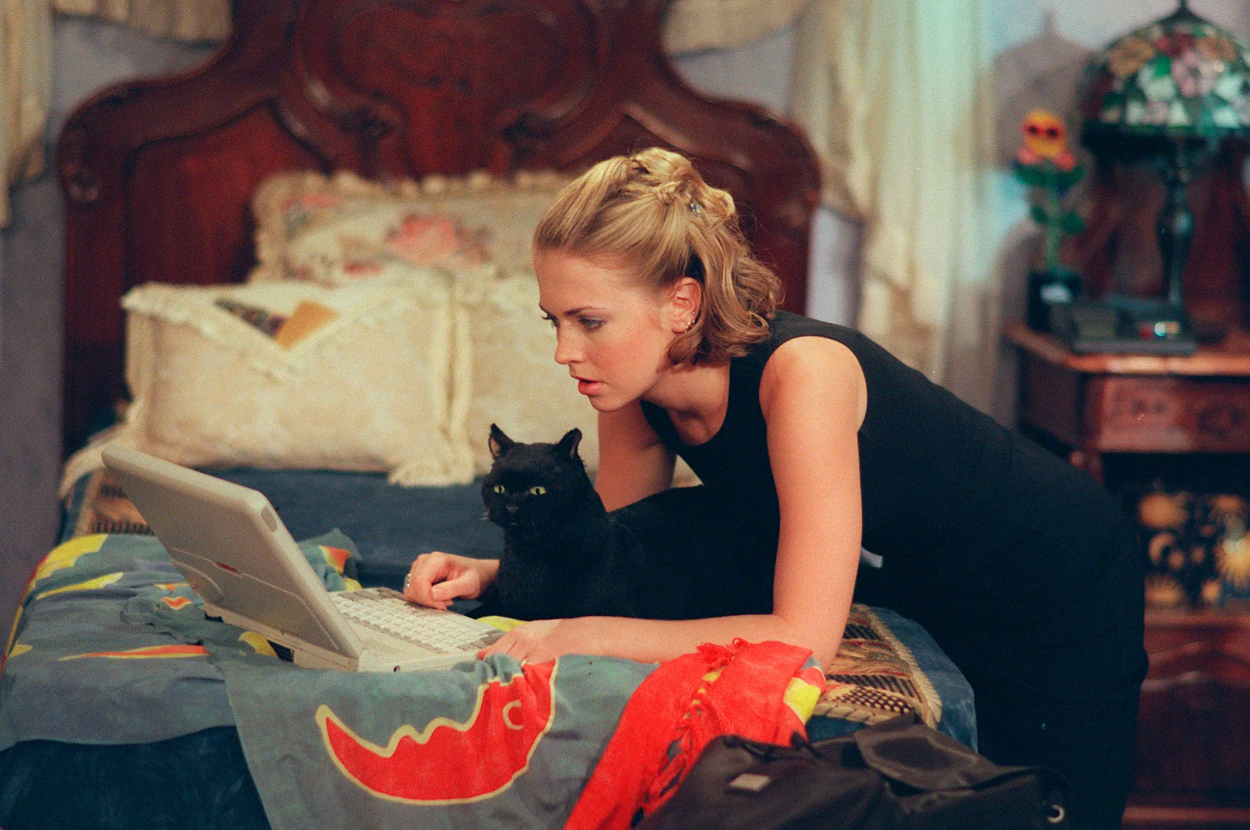 Woman in sleeveless top using laptop with black cat beside her, indoors
