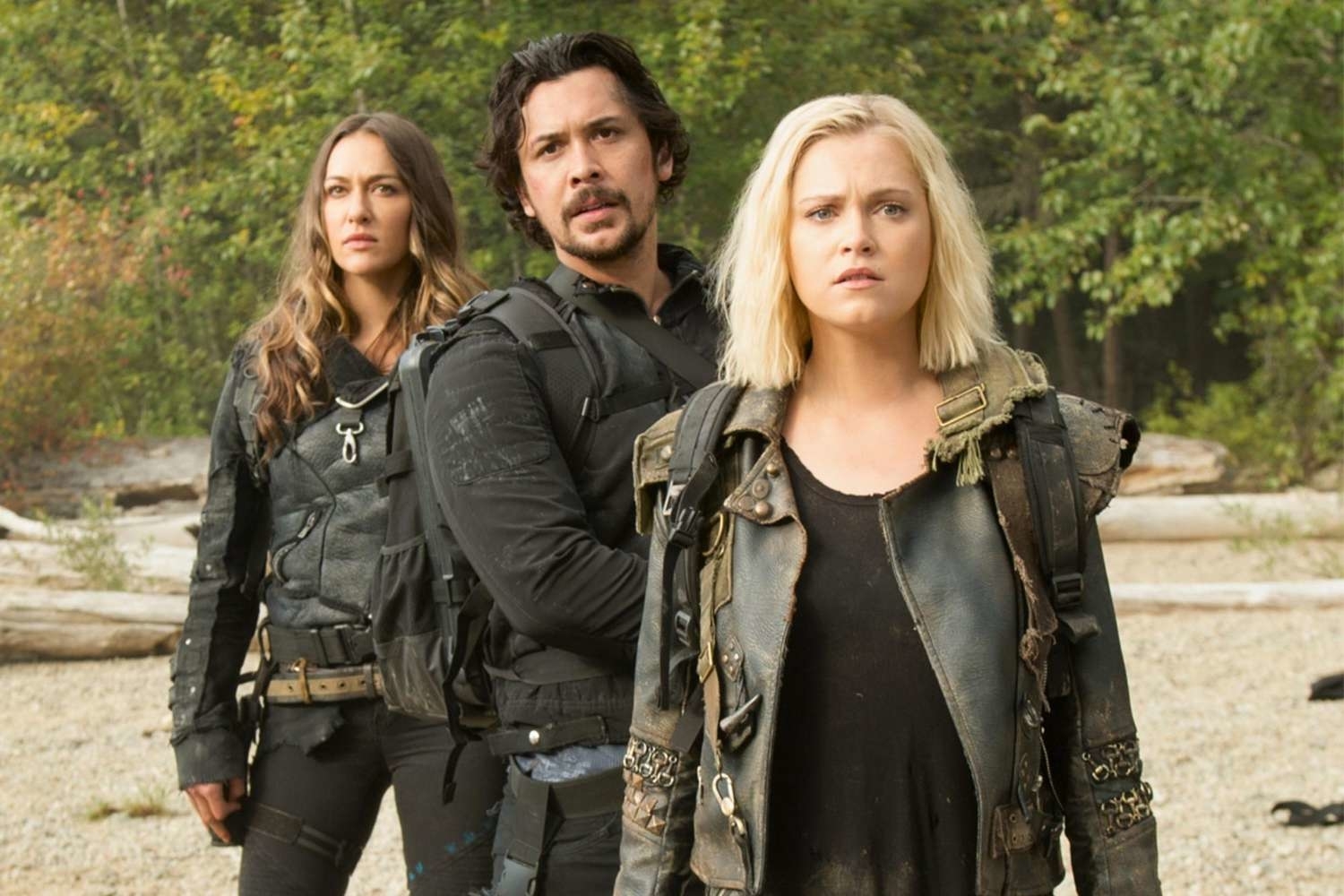 Three actors from the TV show &quot;The 100&quot; standing in a forest, dressed in post-apocalyptic attire