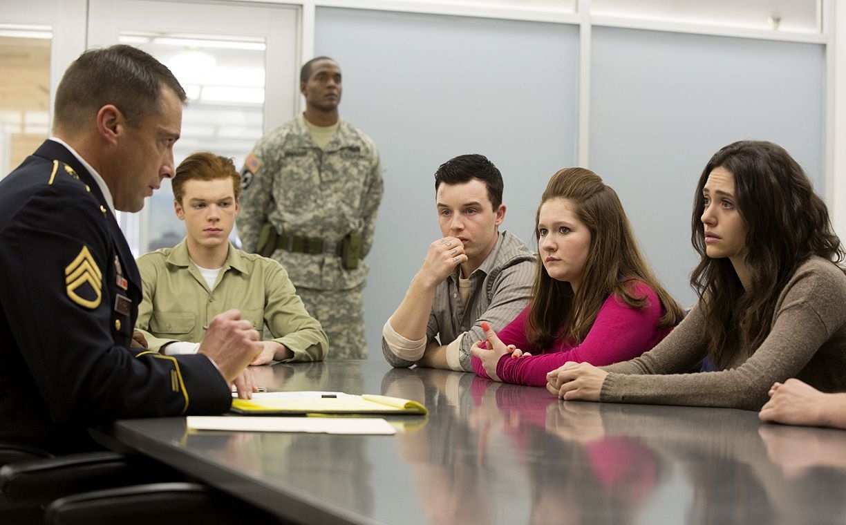 A military officer speaks to four young adults sitting across a table in a briefing room