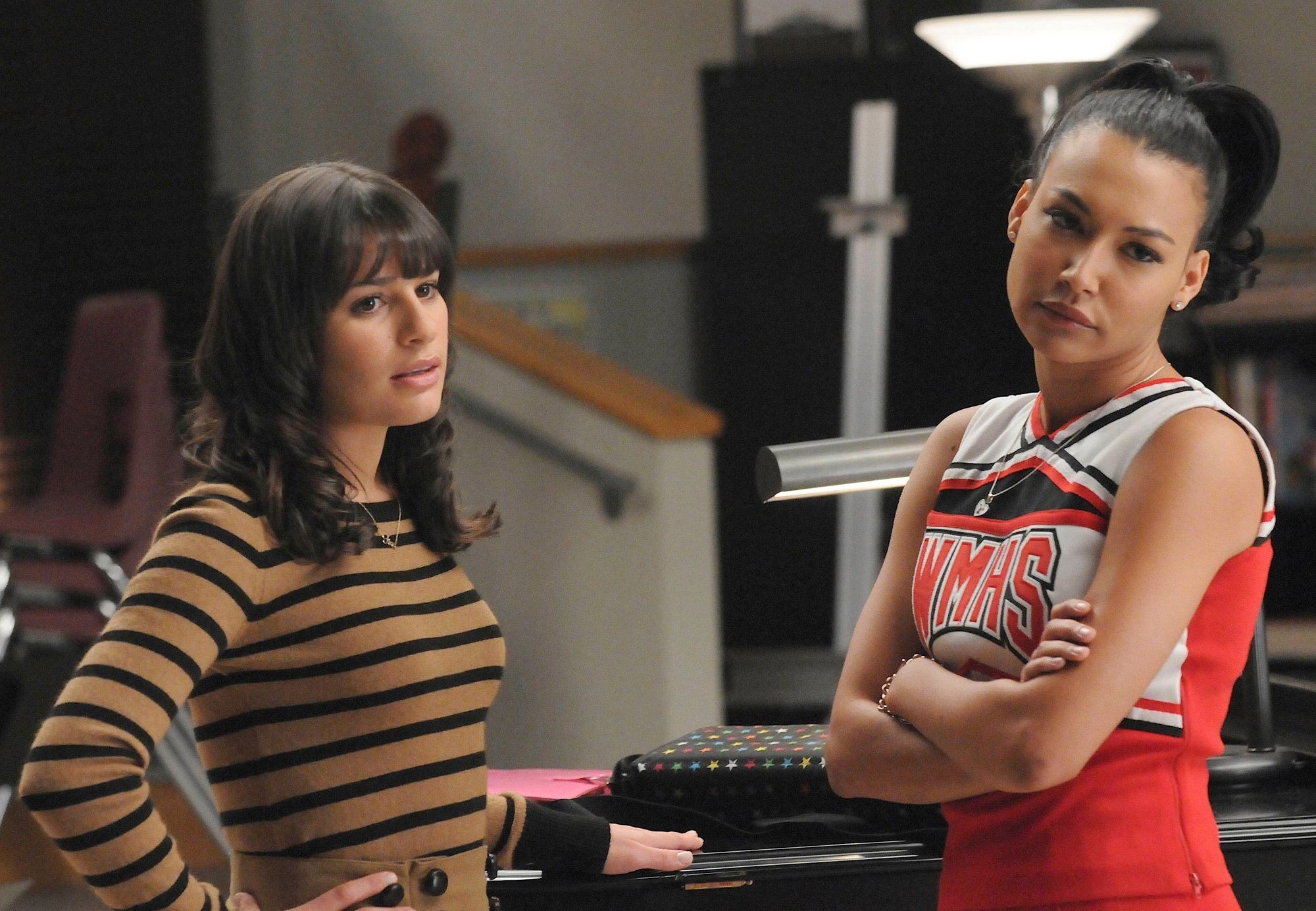 Two characters from Glee, Rachel in a striped sweater and Santana in a cheerleader outfit, stand in a library