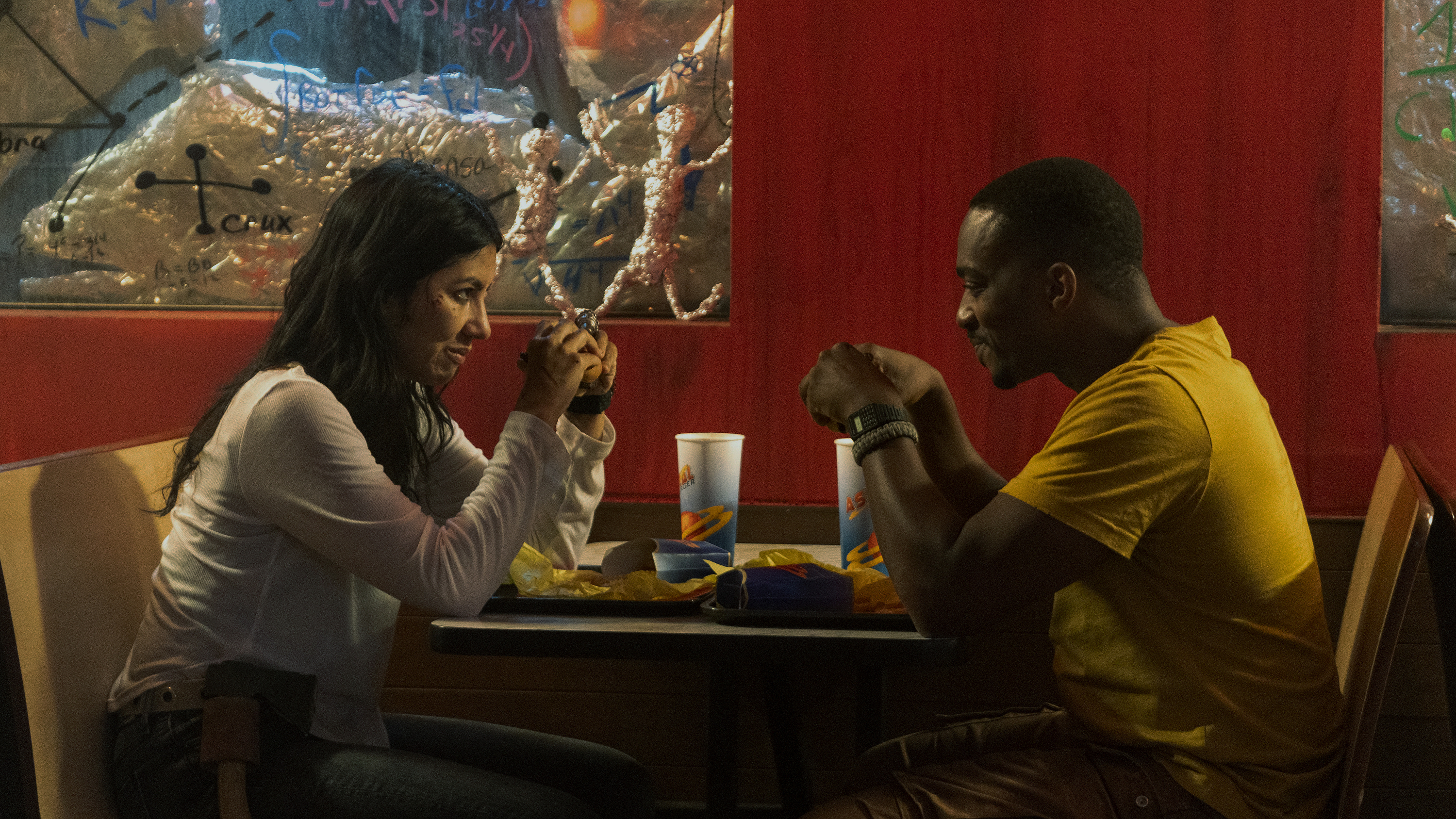 Two characters from a film sit at a diner table, sharing a meal and a conversation