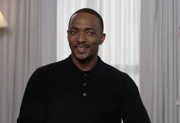 Anthony Mackie smiling at the camera, wearing a black shirt, indoors with curtains in the background