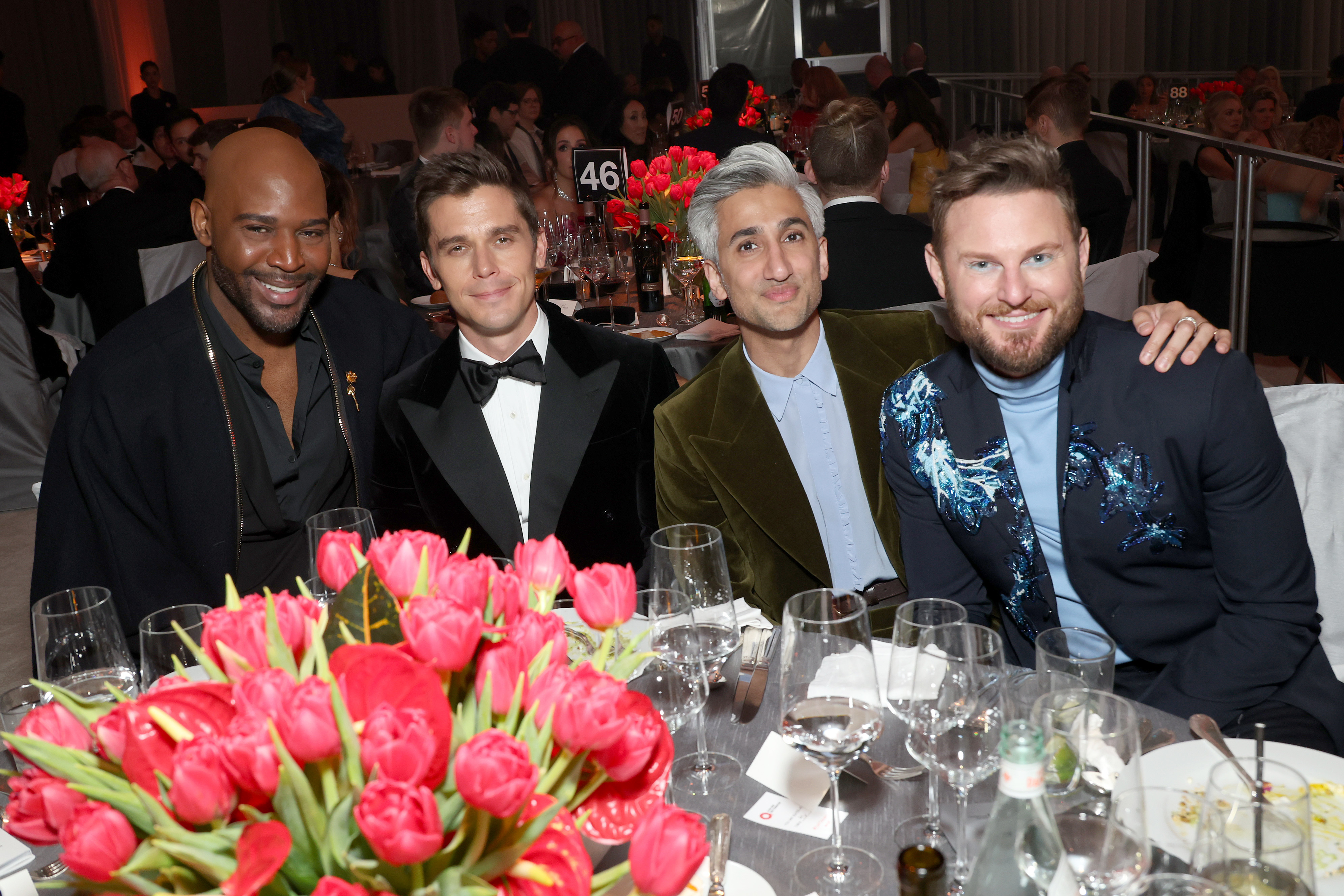 The cast of Queer Eye at an event, seated at a table with a floral centerpiece, smiling for the camera; Tan has his arm around Bobby&#x27;s shoulder