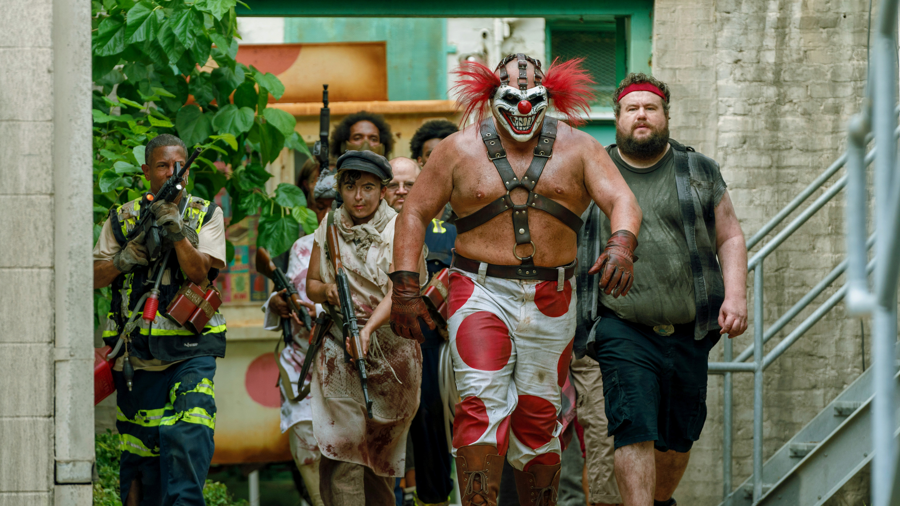 Group of individuals in various costumes and attire walking down an urban alley, led by a person with dramatic makeup and warrior-like outfit