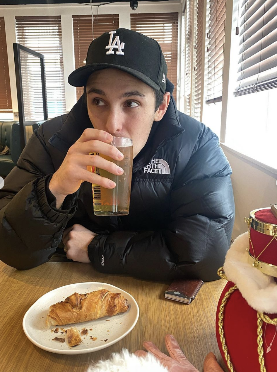 Dalton sipping a drink with a pastry on the table, wearing a baseball cap and jacket