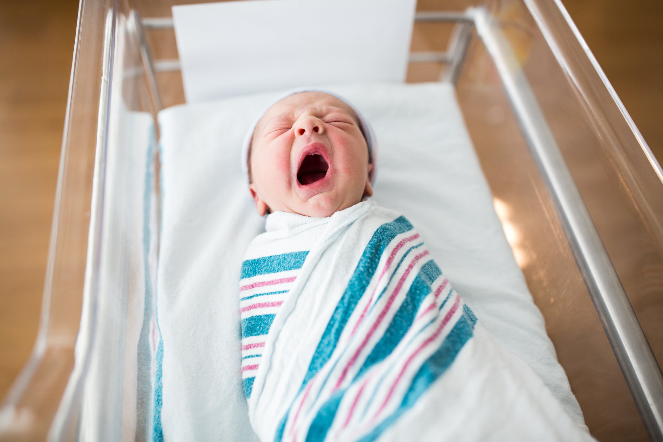 Newborn baby yawning while swaddled in a striped blanket in a hospital bassinet
