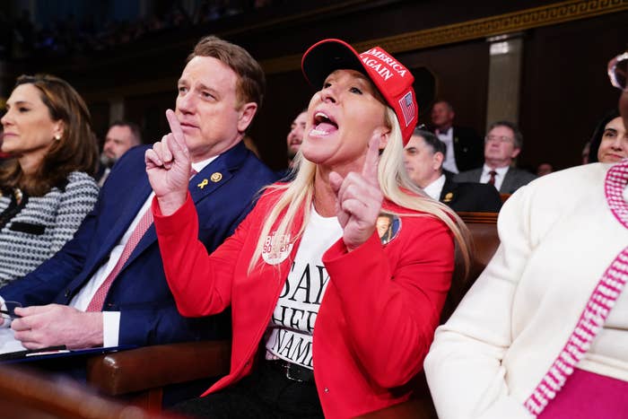 Woman in "Make America Great Again" hat gesturing, seated next to a man in a suit at a formal event