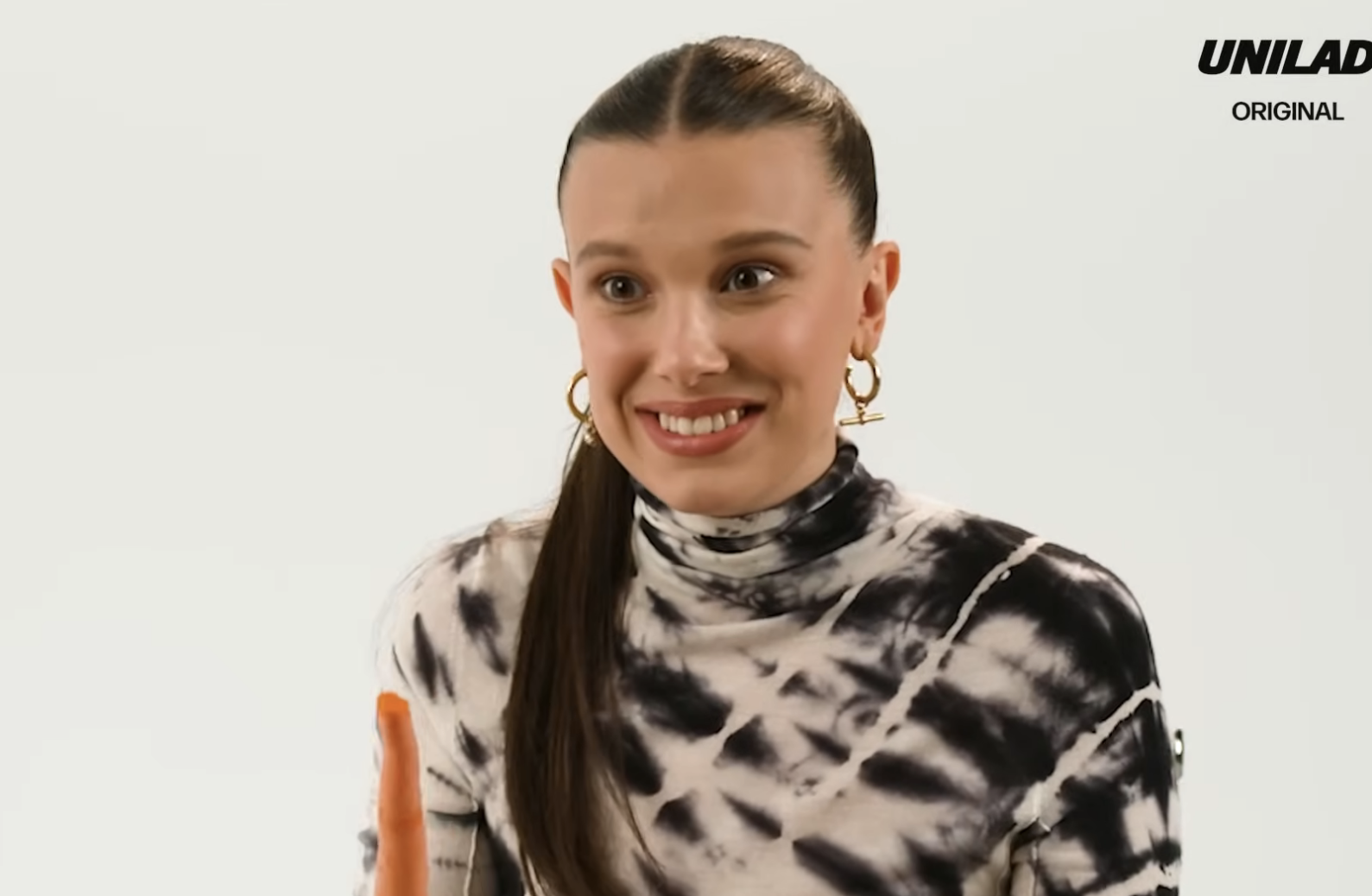 Millie Bobby Brown smiling in a spotted top, holding a carrot