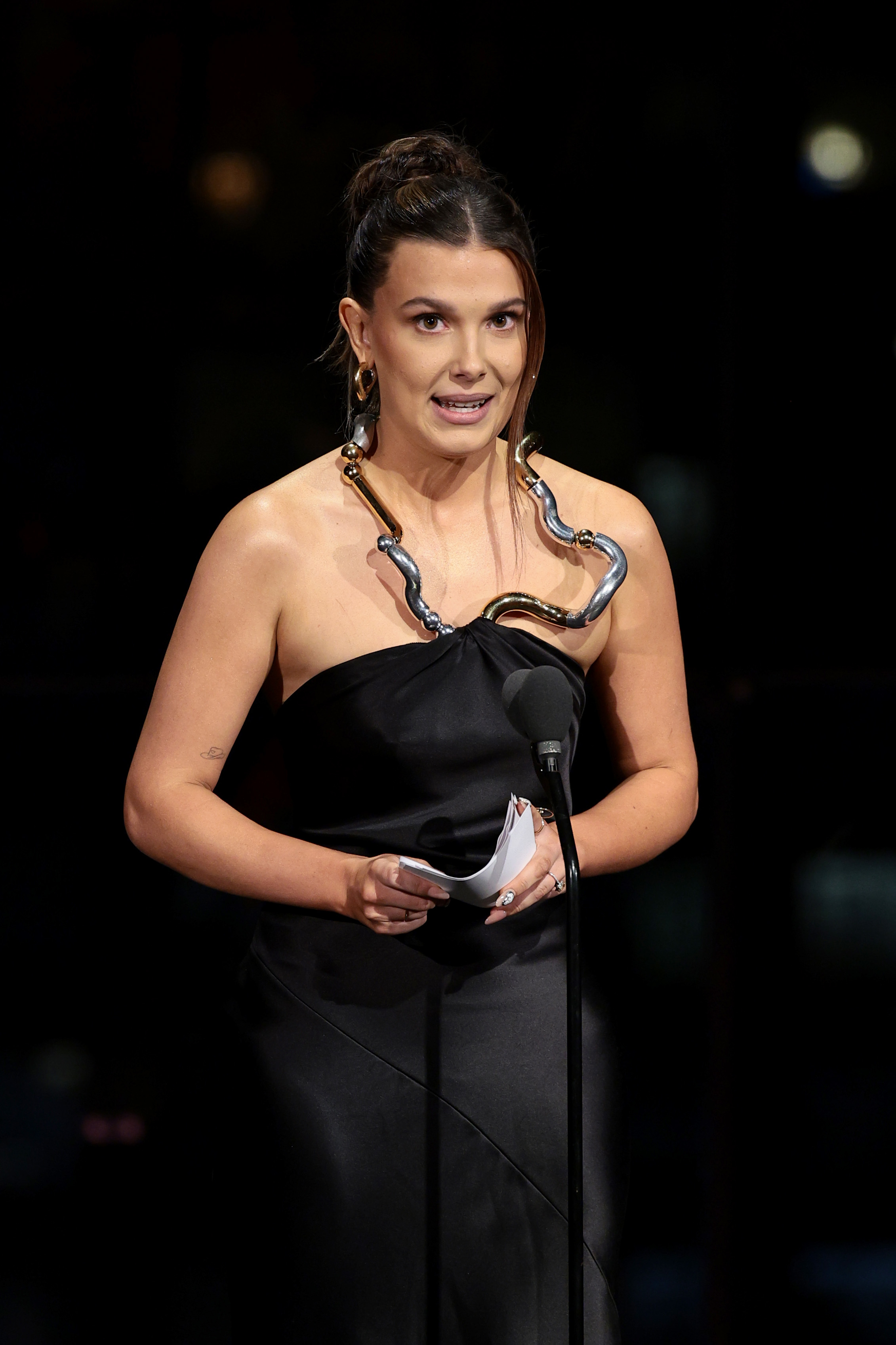 Millie onstage in front of a microphone, wearing a dress with a metal neckline, holding notes