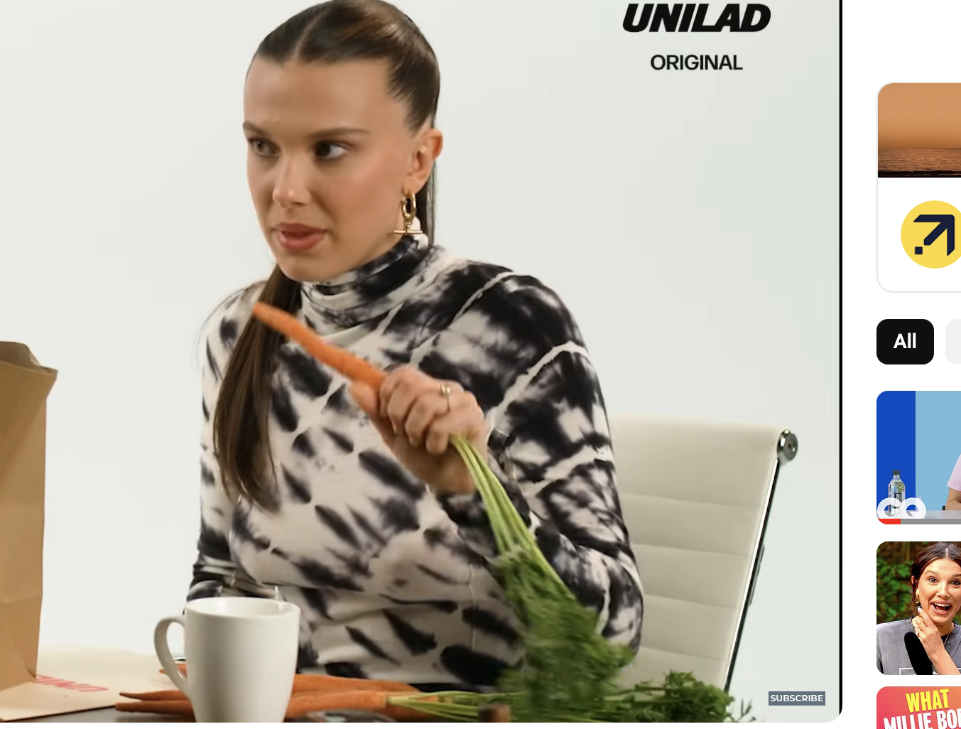 Millie Bobby Brown in a still from a video interview, is seated and holding a carrot with the long stem still attached. On the table in front of her is a paper bag and a mug