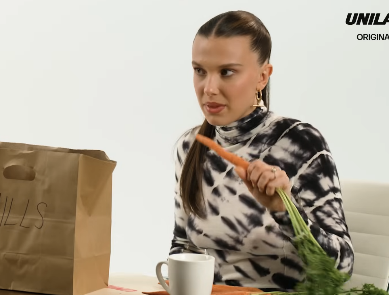 Millie Bobby Brown in a still from a video interview, is seated and holding a carrot with the long stem still attached. On the table in front of her is a paper bag and a mug