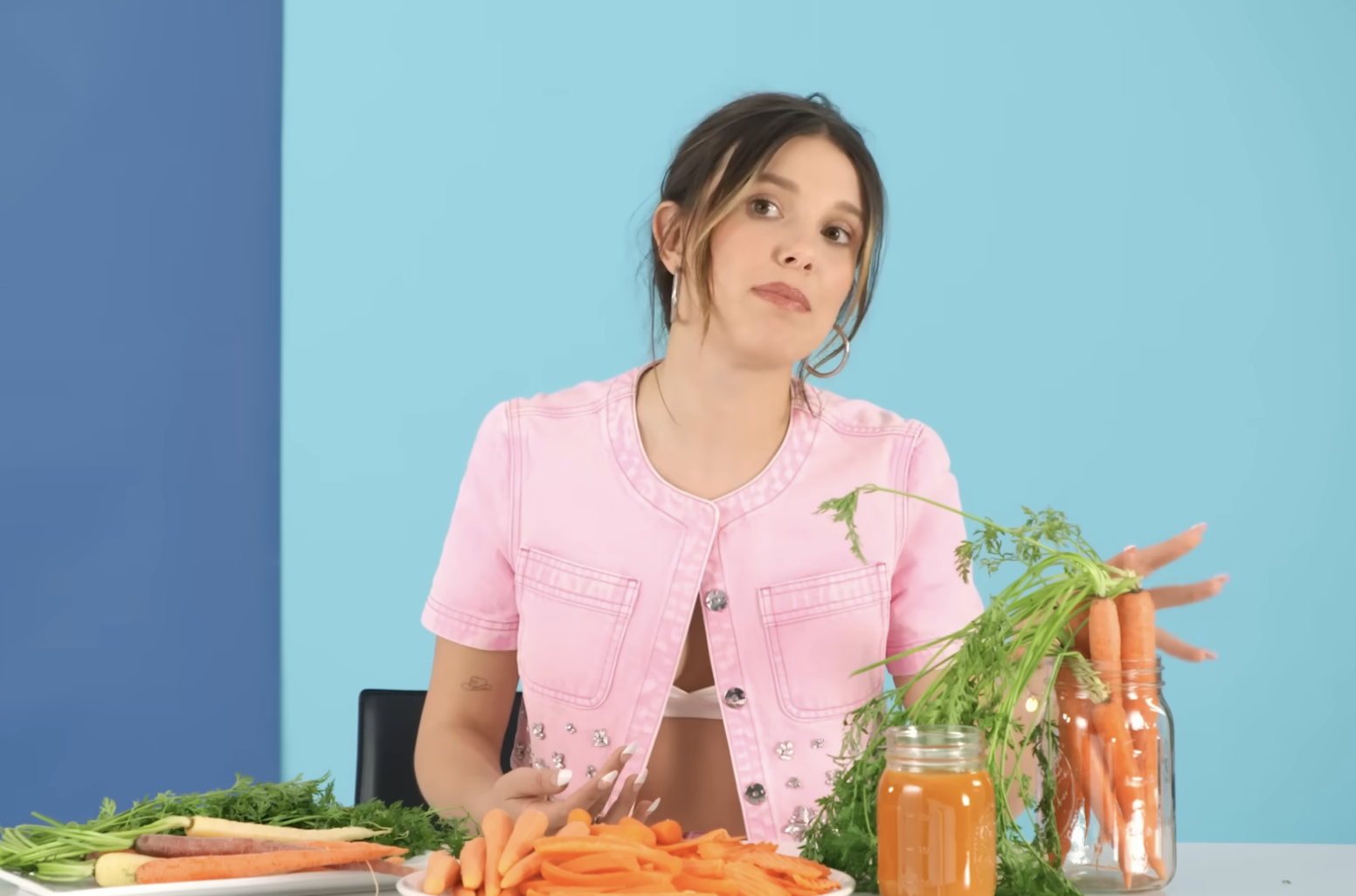 Millie standing behind a table with various items, like a plush toy, snacks, and juice, in a casual setting