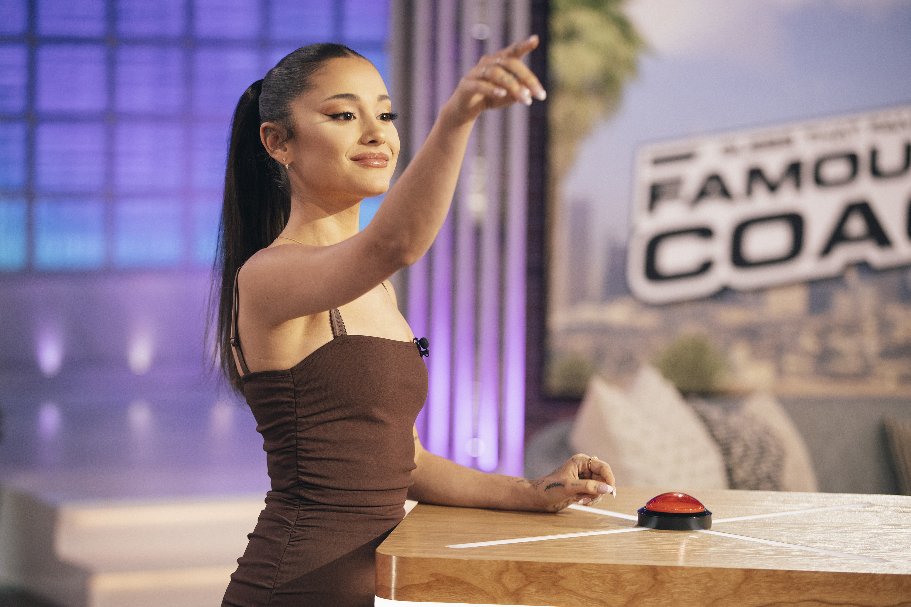 ariana on a game show set, wearing a dress and gesturing with her arm with buzzers on the table
