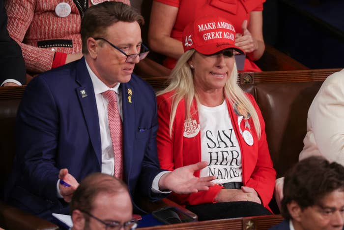Two individuals at an event, one wearing a hat with political slogan, the other in a shirt with a text message