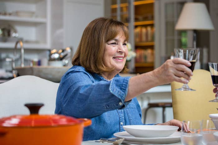 Ina Garten at table smiling, extending a wine glass, with dinnerware and a pot in front