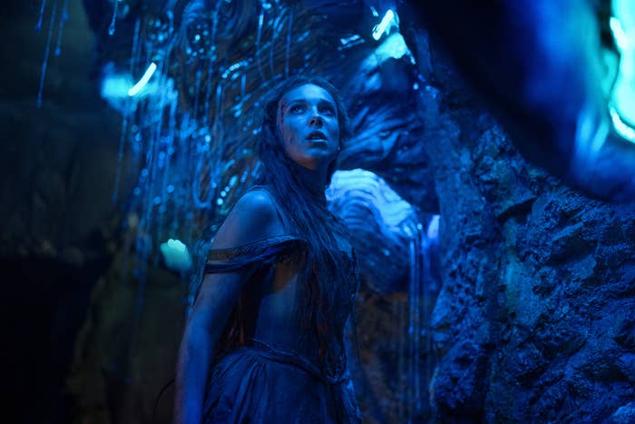 A scene from Damsel showing Millie&#x27;s character in tattered attire, looking intently, surrounded by cave-like eerie blue light