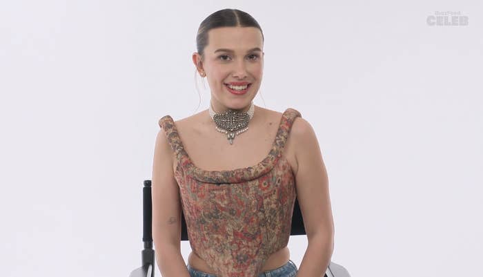 Millie smiling as she sits in a chair. She is wearing a patterned corset top and a choker necklace