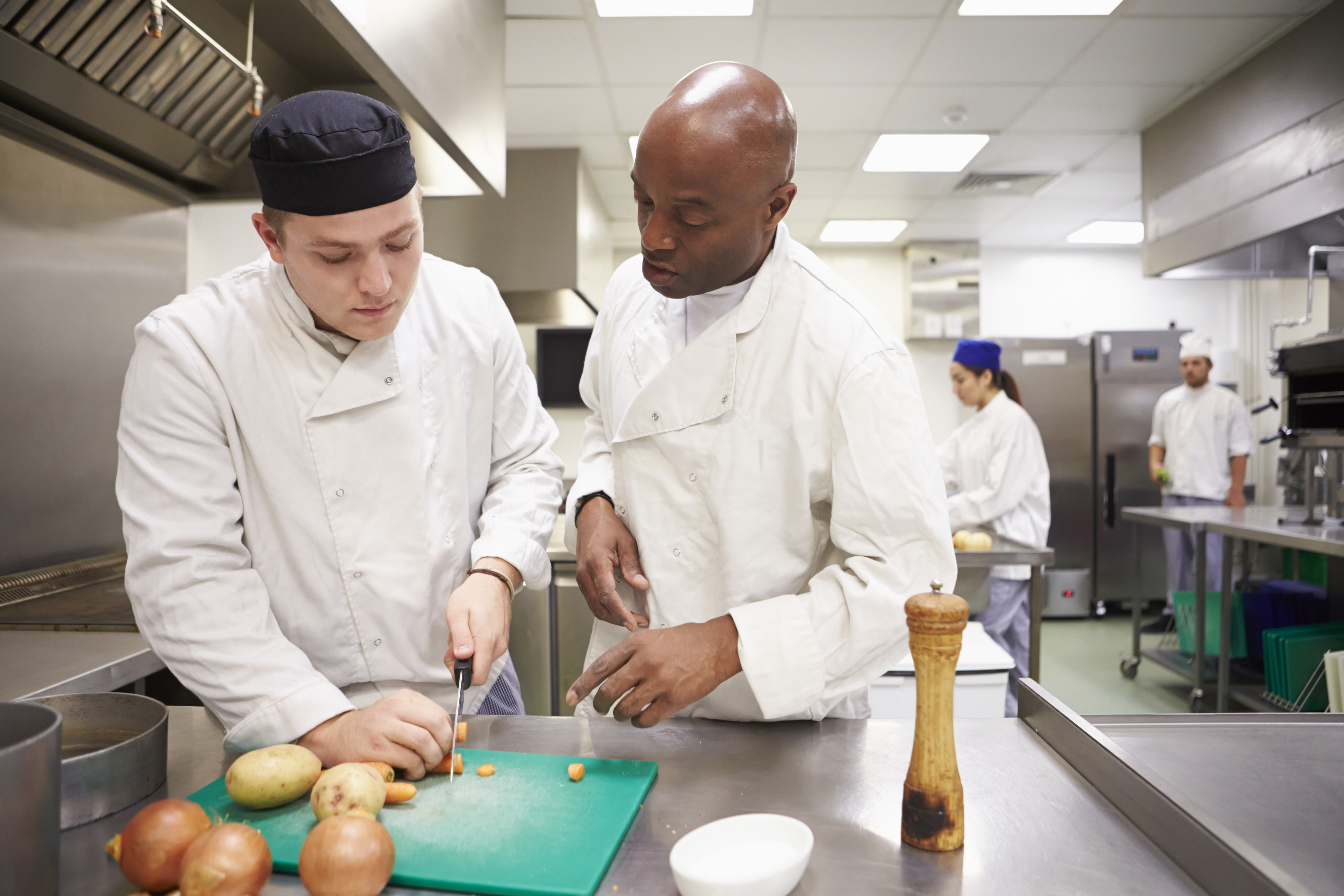 Two chefs, one instructing the other, preparing food in a commercial kitchen