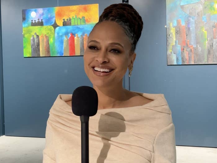 Woman with updo hairstyle smiling, wearing off-shoulder top, standing at a microphone with abstract paintings in the background