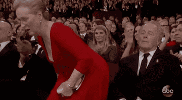 Mujer en traje largo levantándose de su asiento en un evento de gala con asistentes sentados alrededor