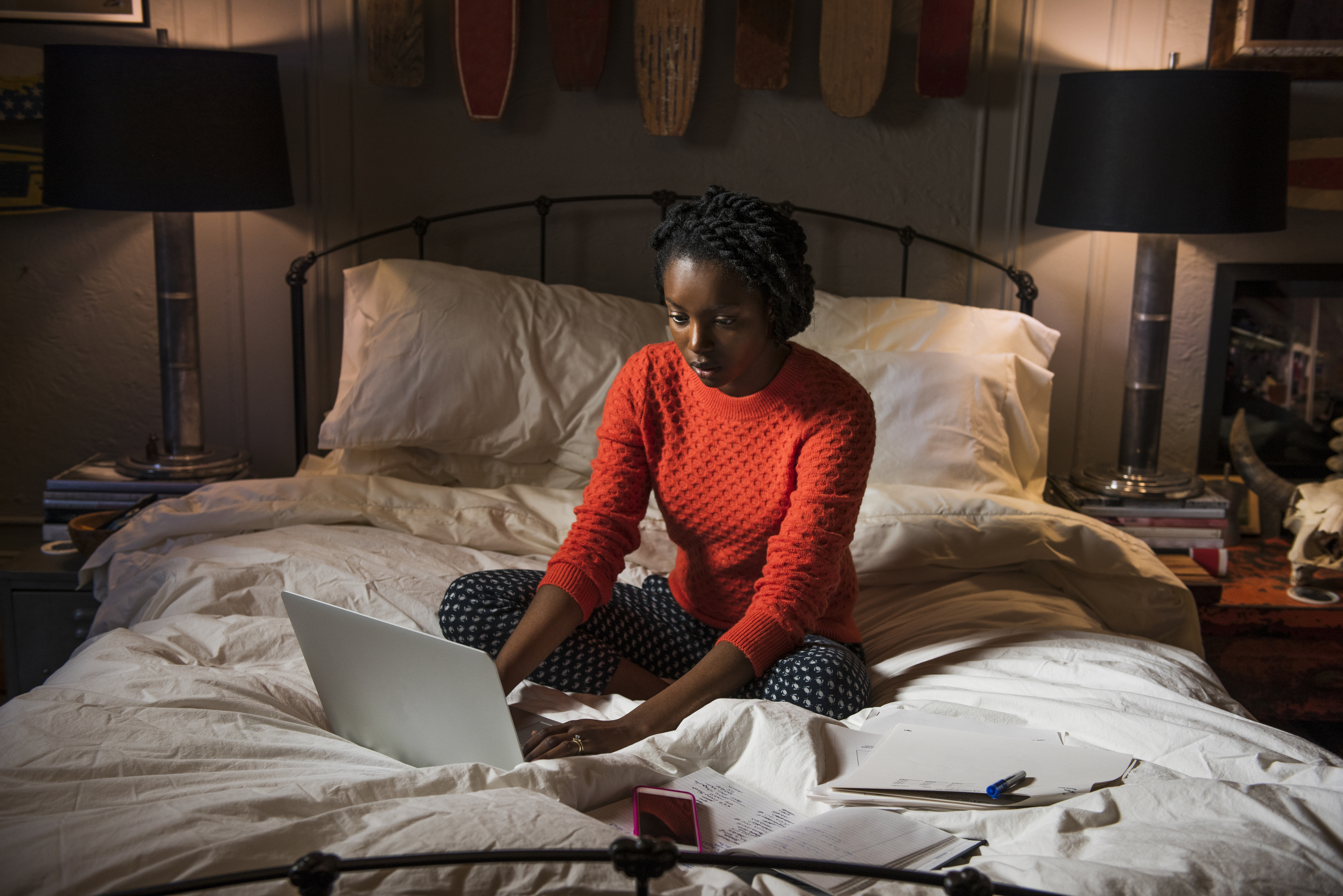 A woman sitting on a  bed with a laptop and papers, looking focused, possibly working or studying late at night