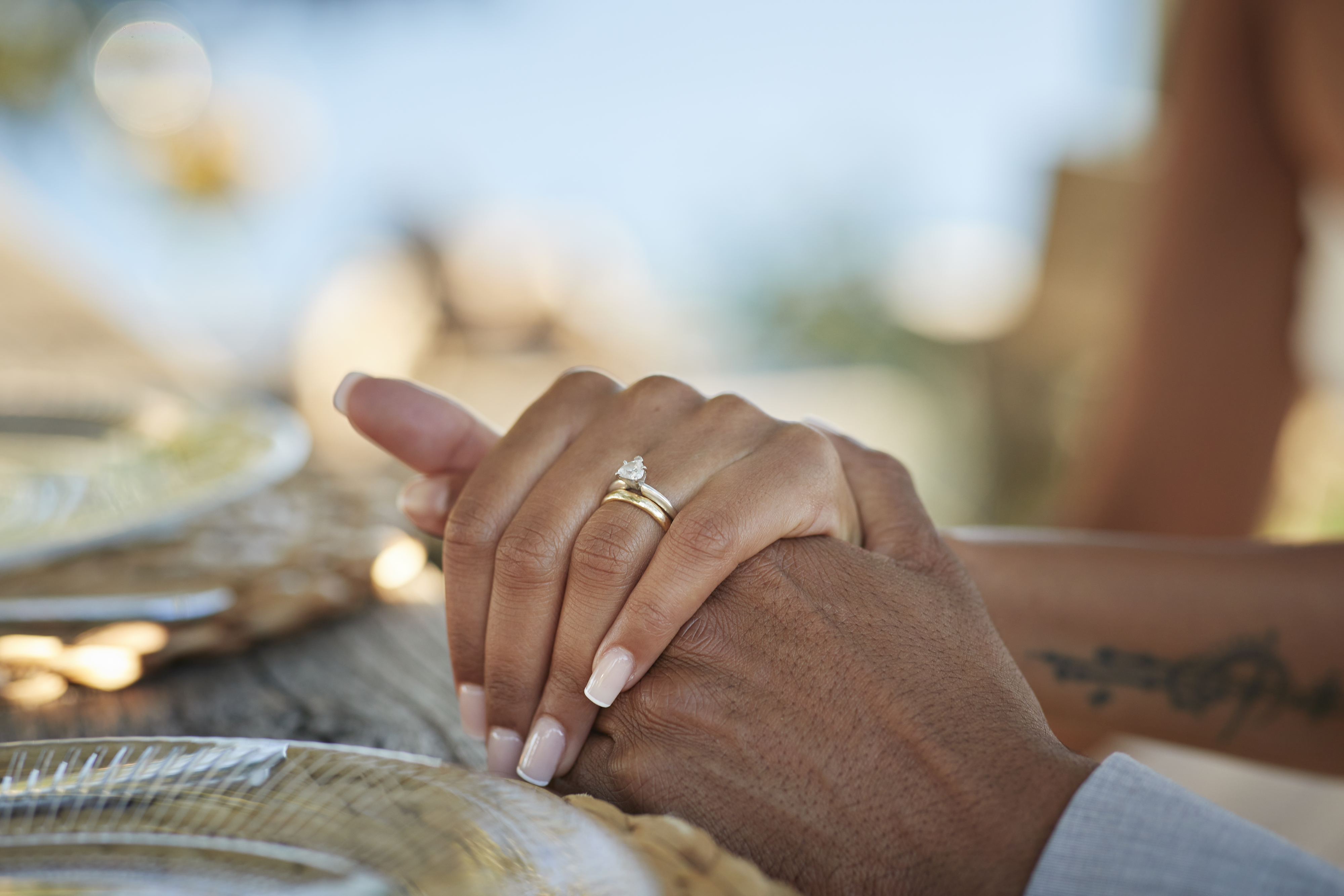 Close-up of a person&#x27;s hands clasped on a table, one with a tattoo and wearing a ring