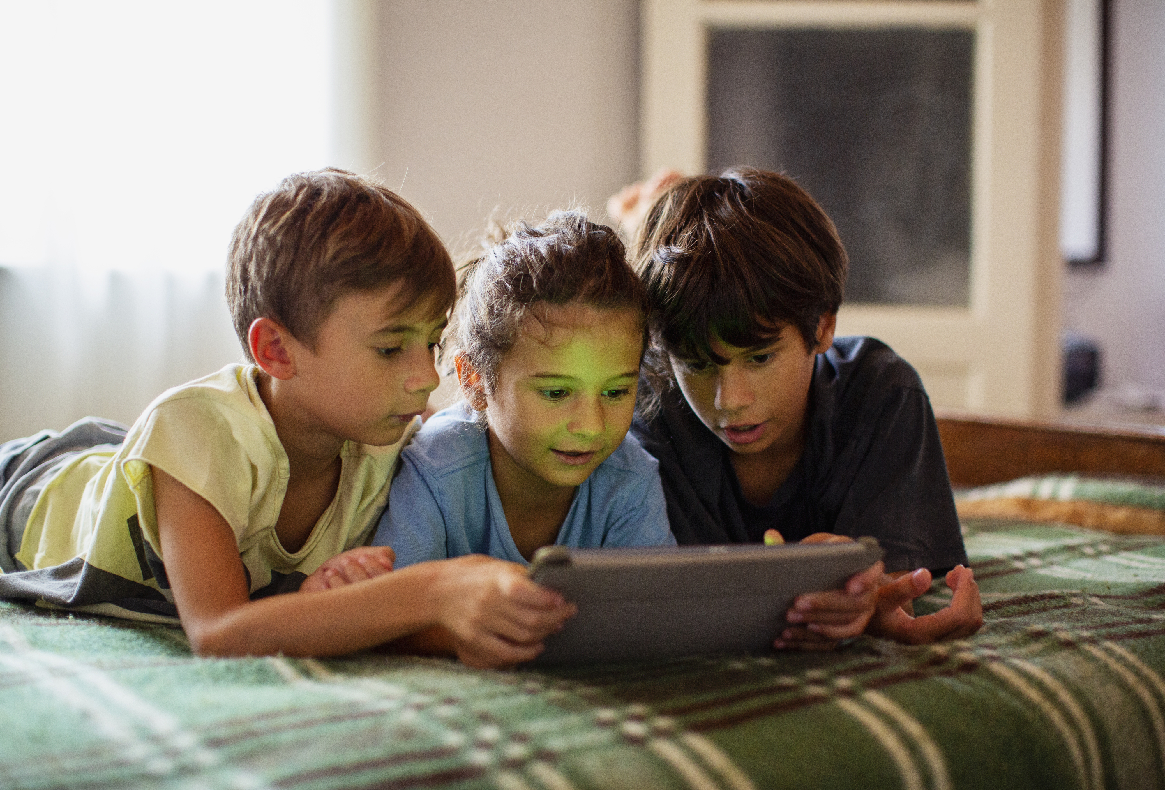 Three children lying on a bed looking at a tablet together