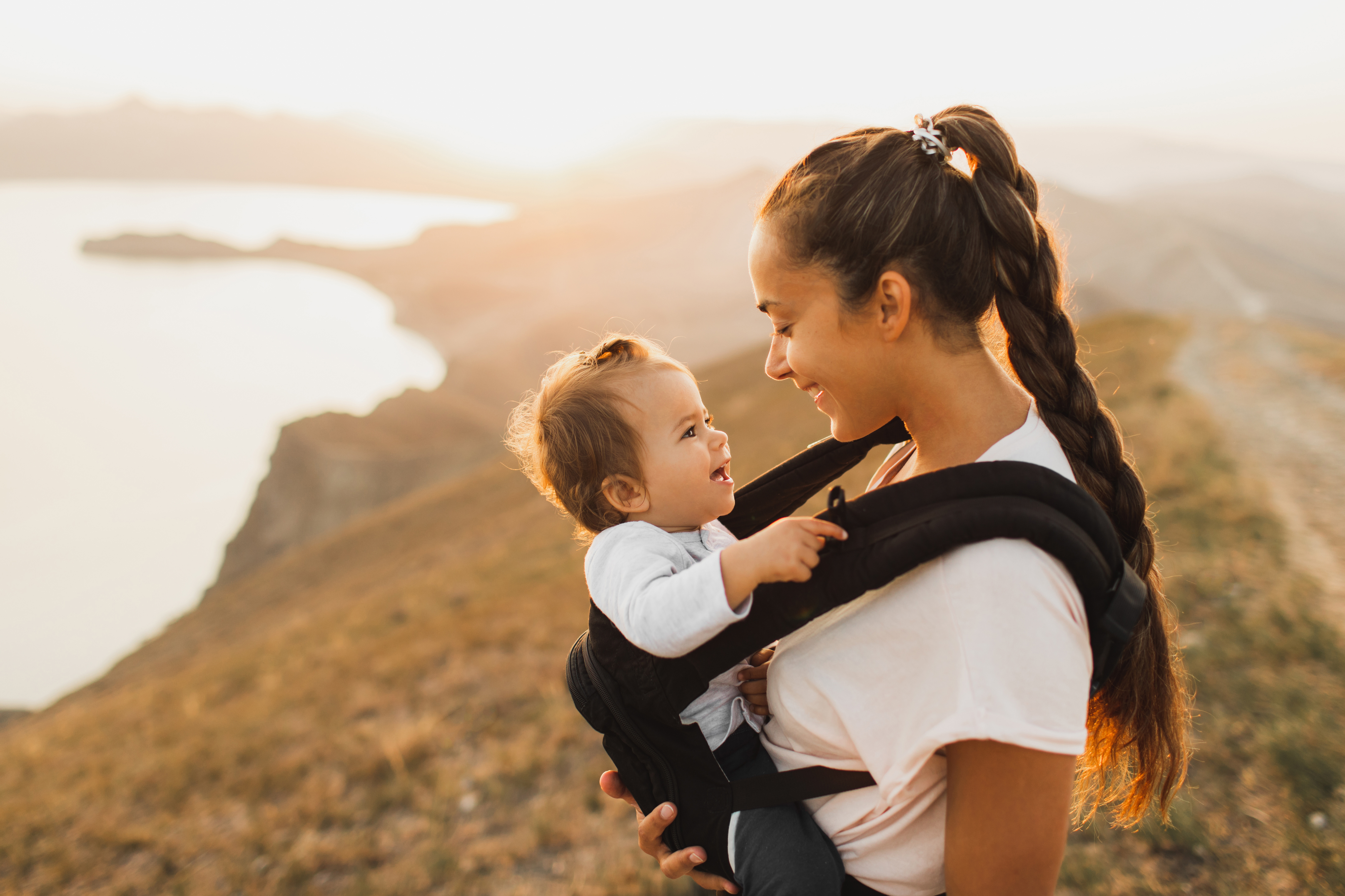 Woman with a baby in a carrier, both smiling, on a hillside at sunset
