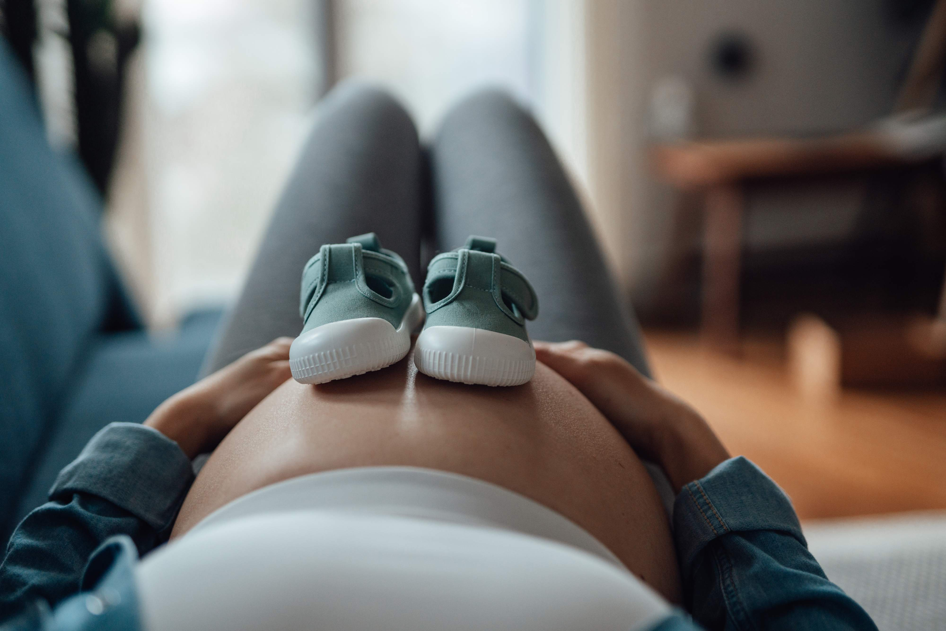 Pregnant person resting on a sofa with baby shoes on their belly