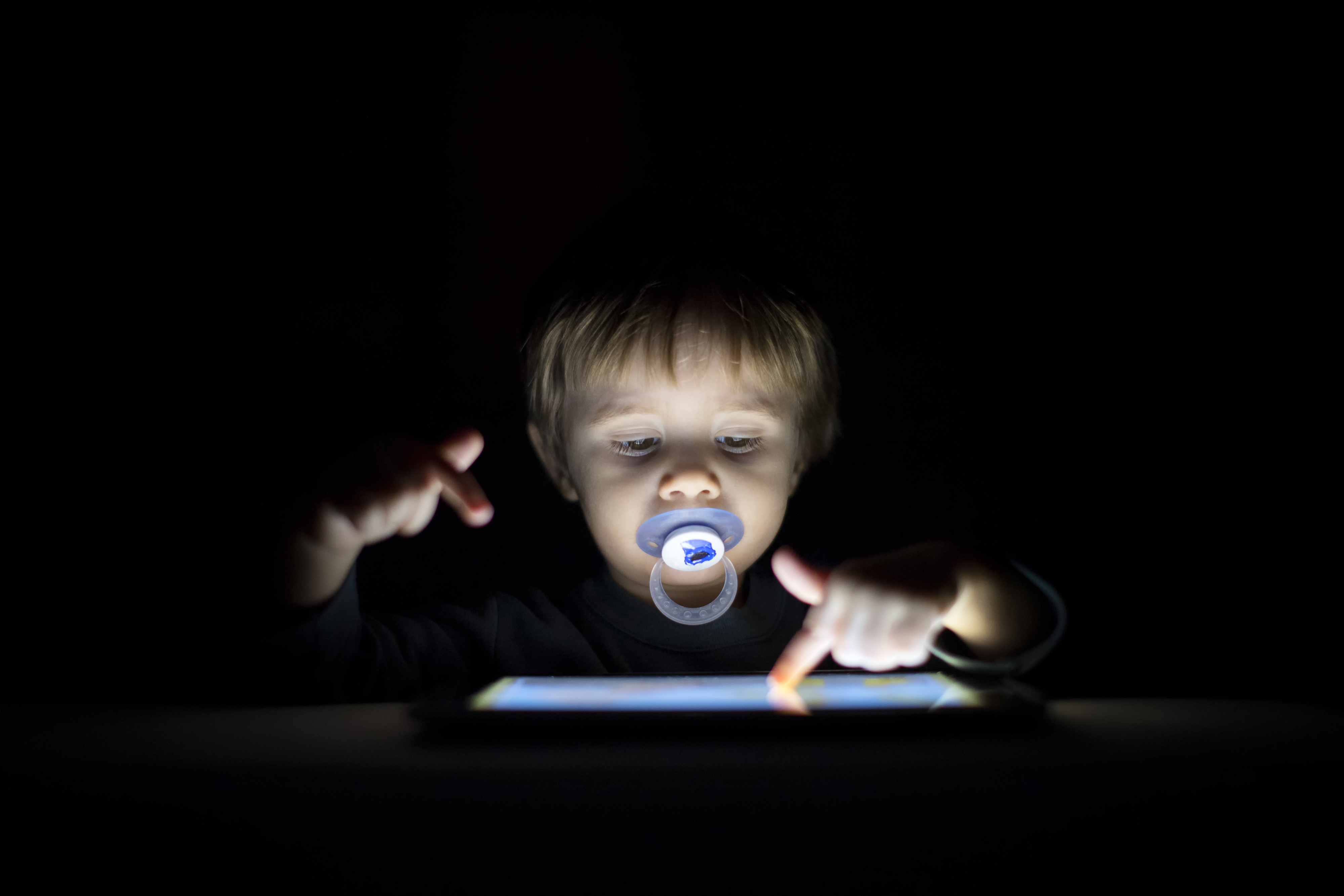 A toddler with a pacifier in their mouth using a tablet in a dark room with the tablet illuminating their face