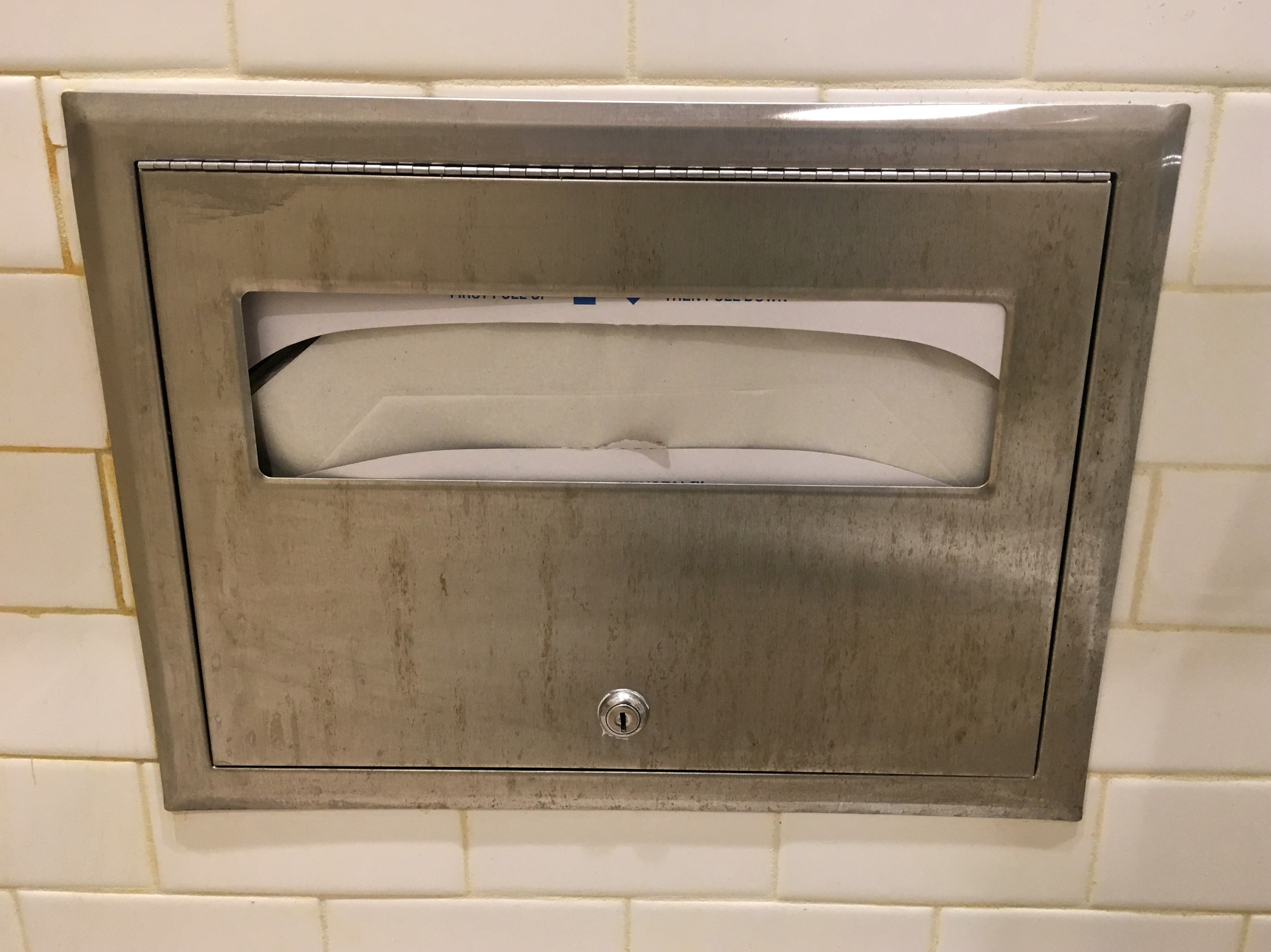 A metal paper towel dispenser embedded in a tiled wall, filled with paper towels, with a small viewing window