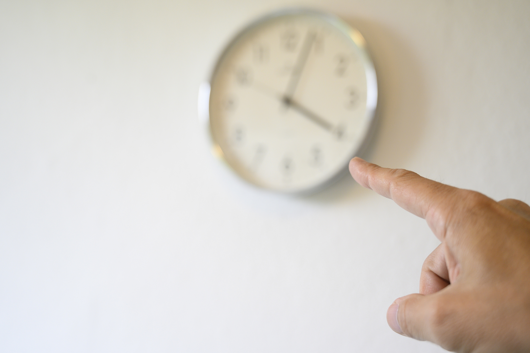 A person&#x27;s index finger pointing towards a wall-mounted clock indicating a time around 10:10