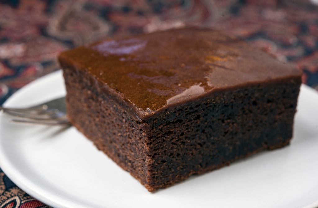 A moist chocolate cake slice on a white plate with a fork alongside